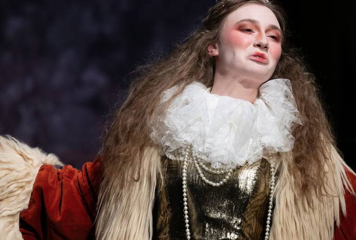 A close-up of a theatrical performer dressed in an elaborate costume. The person wears a rich velvet garment with fur trim, a ruffled lace collar, and a string of pearls. Their makeup is dramatic, with rosy cheeks and pronounced features, giving an aristocratic or regal appearance. Their expression is poised and commanding, and their arms are slightly raised as if delivering a powerful monologue or gesture. The background is dark and muted, emphasizing the performer’s presence.
