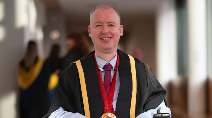 Stephen McGuire, a Paralympic gold medallist, wearing formal graduation attire with gold trim and a medal around his neck, seated in a wheelchair. He is smiling and positioned in a well-lit hallway, with blurred figures of graduates in the background.