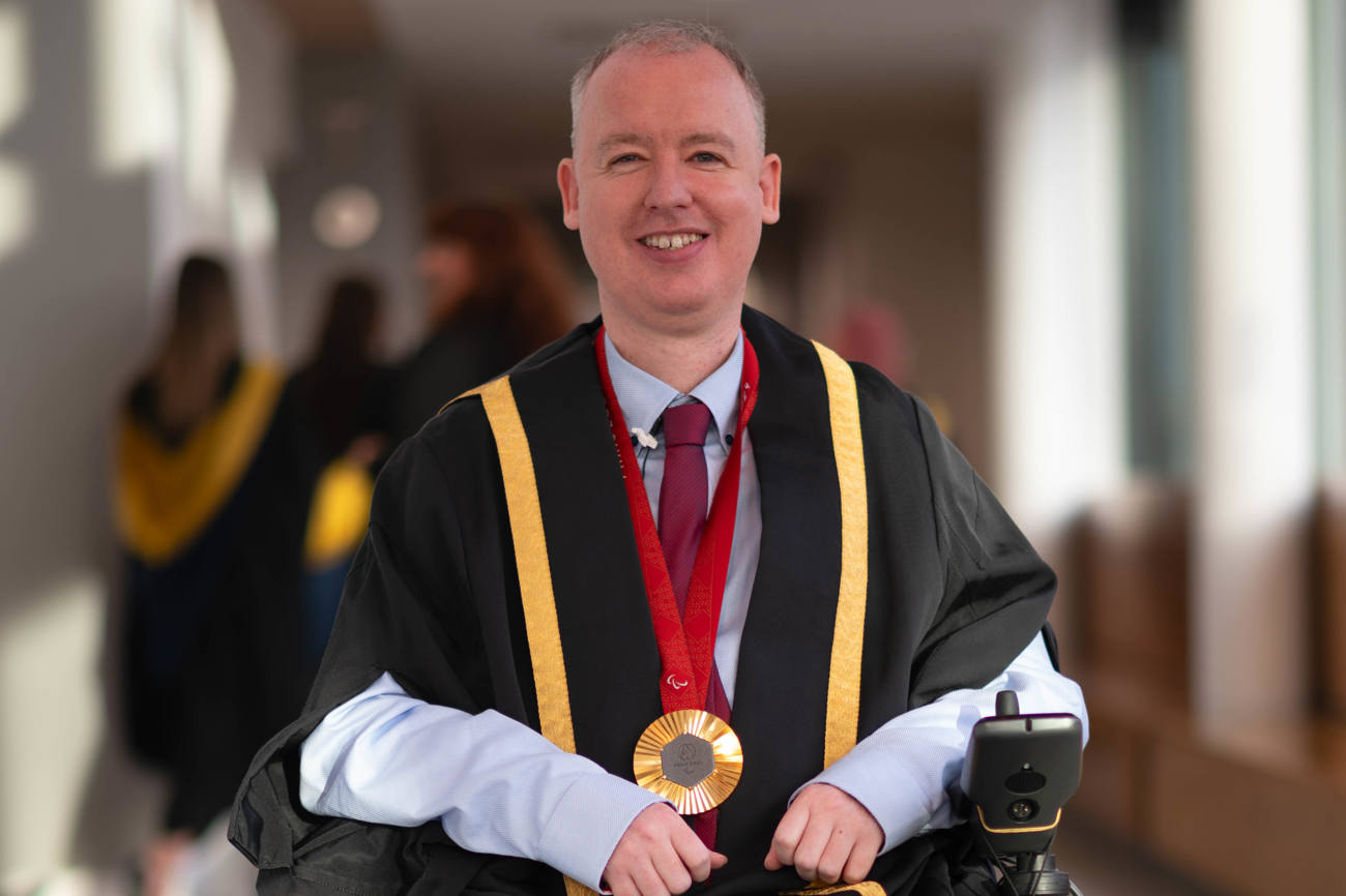 Stephen McGuire, a Paralympic gold medallist, wearing formal graduation attire with gold trim and a medal around his neck, seated in a wheelchair. He is smiling and positioned in a well-lit hallway, with blurred figures of graduates in the background.