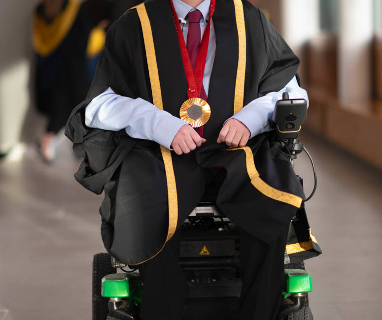 Stephen McGuire, a Paralympic gold medallist, wearing formal graduation attire with gold trim and a medal around his neck, seated in a wheelchair. He is smiling and positioned in a well-lit hallway, with blurred figures of graduates in the background.