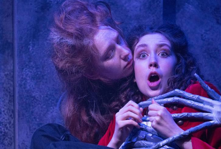 A theatrical scene featuring two individuals in an intense moment. One person, with wild hair and a pale complexion, leans closely toward another who looks startled and wide-eyed. The second person is dressed in a red garment and appears to be gripped by a skeletal, bony hand wrapped around their shoulders, adding to the dramatic and eerie atmosphere. The background is dimly lit with a textured wall, enhancing the moody and suspenseful tone of the scene.