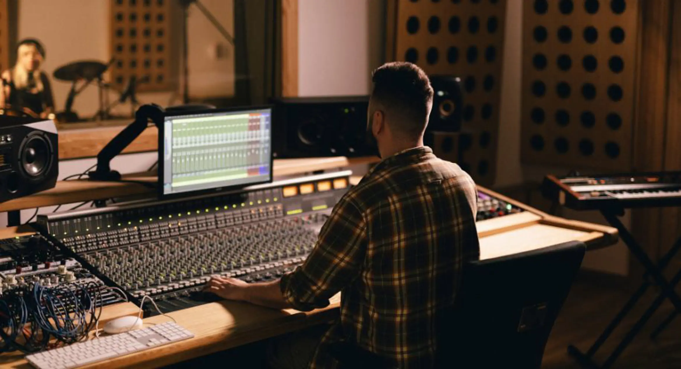 A sound engineer working at a mixing desk in a professional recording studio, with a digital audio workstation displayed on the monitor. The room features studio equipment, soundproofing panels and a musician visible in the background through a recording booth window.