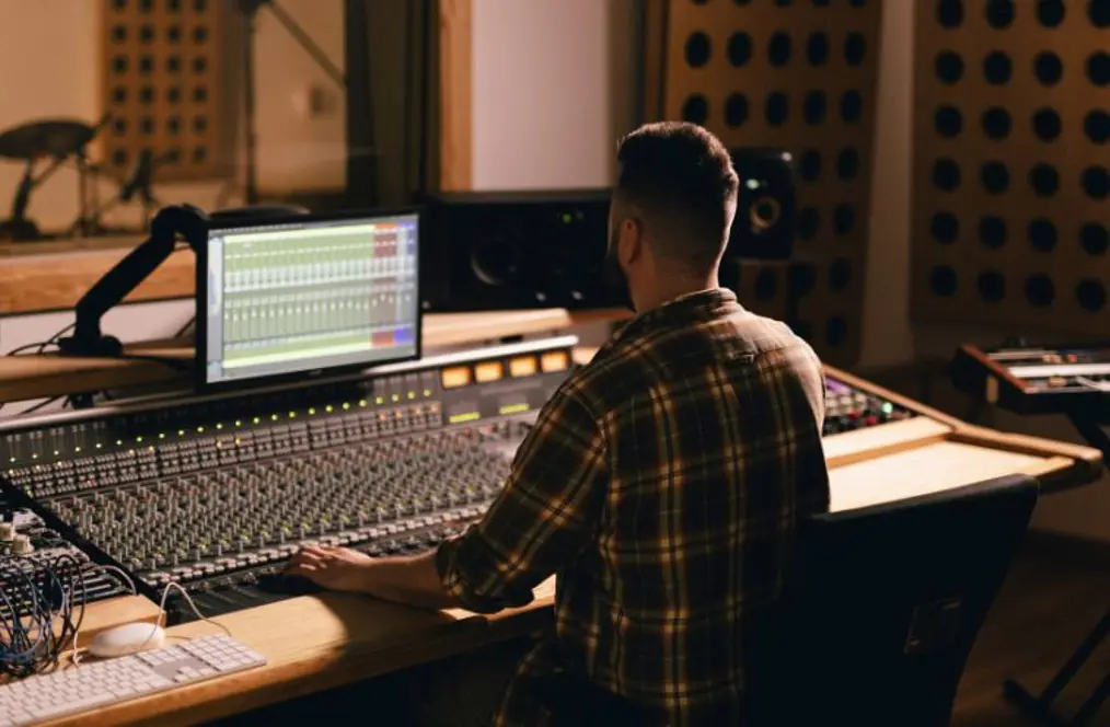A sound engineer working at a mixing desk in a professional recording studio, with a digital audio workstation displayed on the monitor. The room features studio equipment, soundproofing panels and a musician visible in the background through a recording booth window.