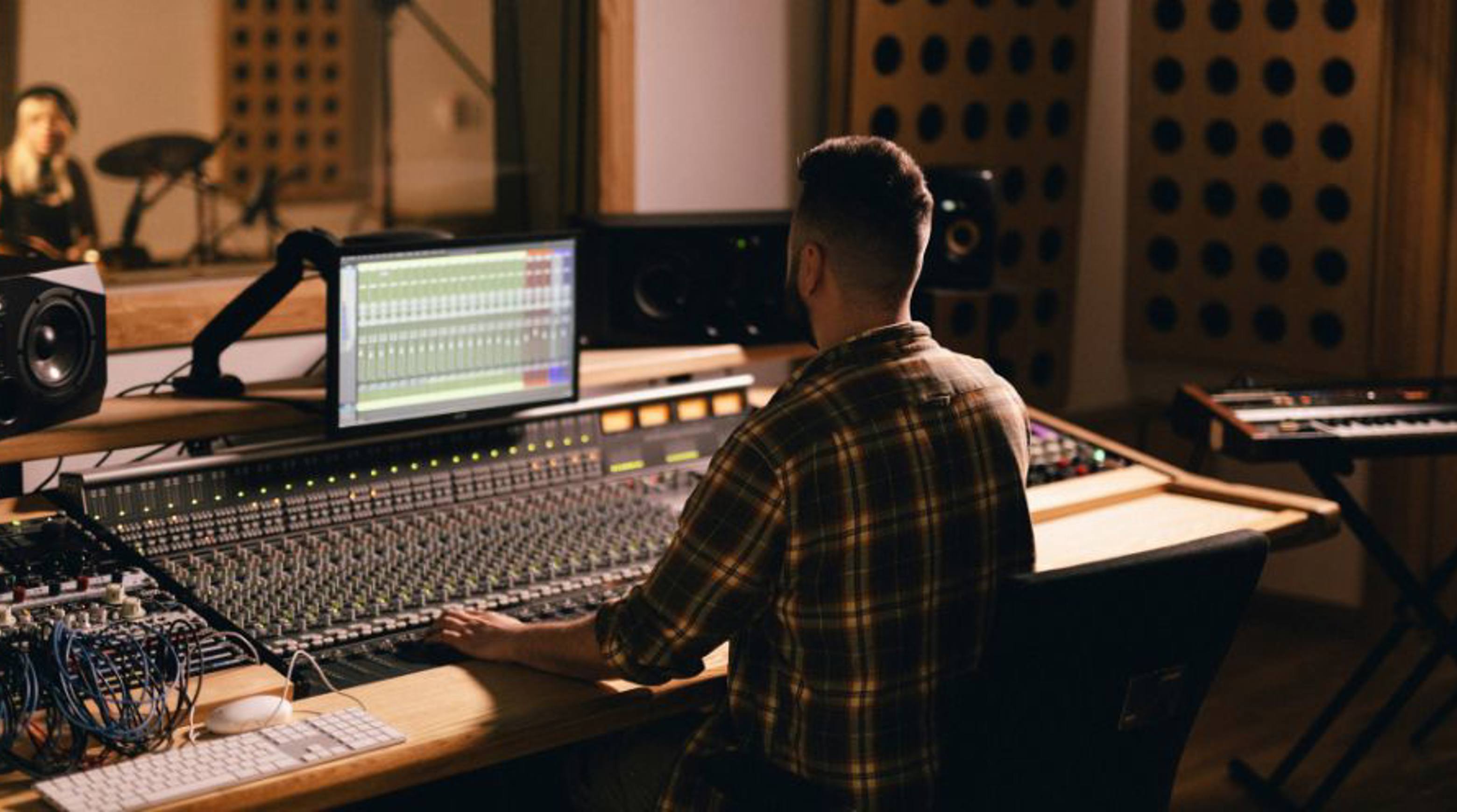 A sound engineer working at a mixing desk in a professional recording studio, with a digital audio workstation displayed on the monitor. The room features studio equipment, soundproofing panels and a musician visible in the background through a recording booth window.