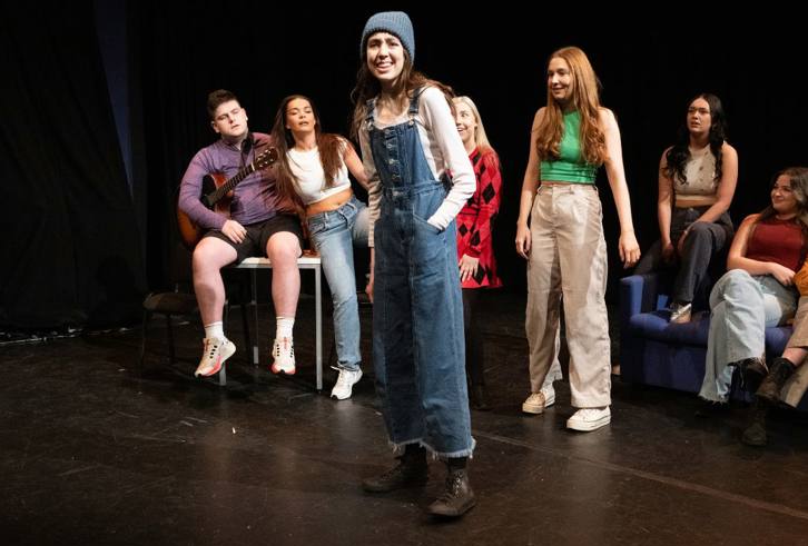 A group of young people is on stage, participating in a theatrical performance. In the foreground, a person in denim overalls, a white long-sleeve shirt, and a beanie stands confidently, engaging with the audience or cast. Behind them, another individual plays a guitar, while others are seated and standing, watching and smiling, creating a lively and supportive atmosphere. The setting is casual, with a dark backdrop, emphasizing the stage's focus on the performers.