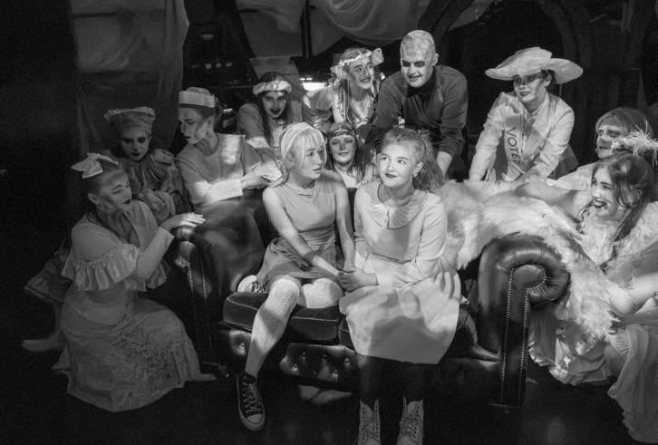 A black-and-white photo depicting a group of people in theatrical costumes. They are gathered around two individuals seated on a couch. The scene has a dramatic and vintage feel, with each person wearing distinct outfits, including period dresses, hats, and accessories. Some have theatrical makeup, adding to the dramatic atmosphere. The lighting highlights the faces and expressions, emphasising the emotional tone of the gathering.