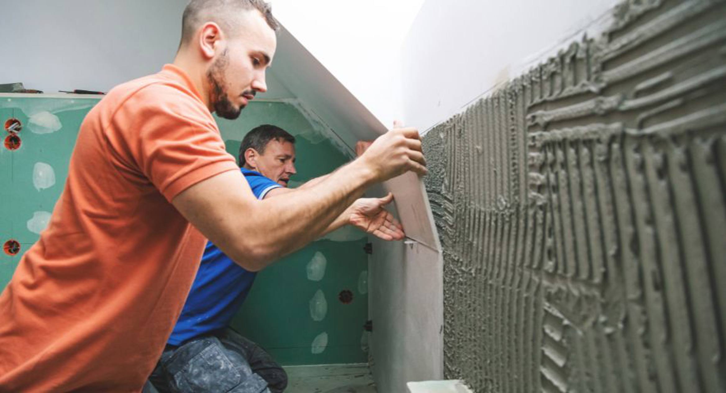 Two men tiling a wall in a room under construction. The man in the foreground is applying adhesive to the wall with a notched trowel, while the man in the background prepares to assist, highlighting teamwork in a hands-on construction or trade setting.