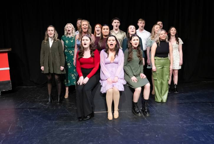 A group of performers is standing in formation on a stage, singing together in unison. They are dressed in a mix of casual and semi-formal attire, featuring various shades of green, red, and neutral tones. The performers have expressive faces, emphasising the emotional intensity of the song. The background is simple with a dark curtain, keeping the focus on the group. The scene suggests a theatrical or musical performance, highlighting teamwork and harmony.